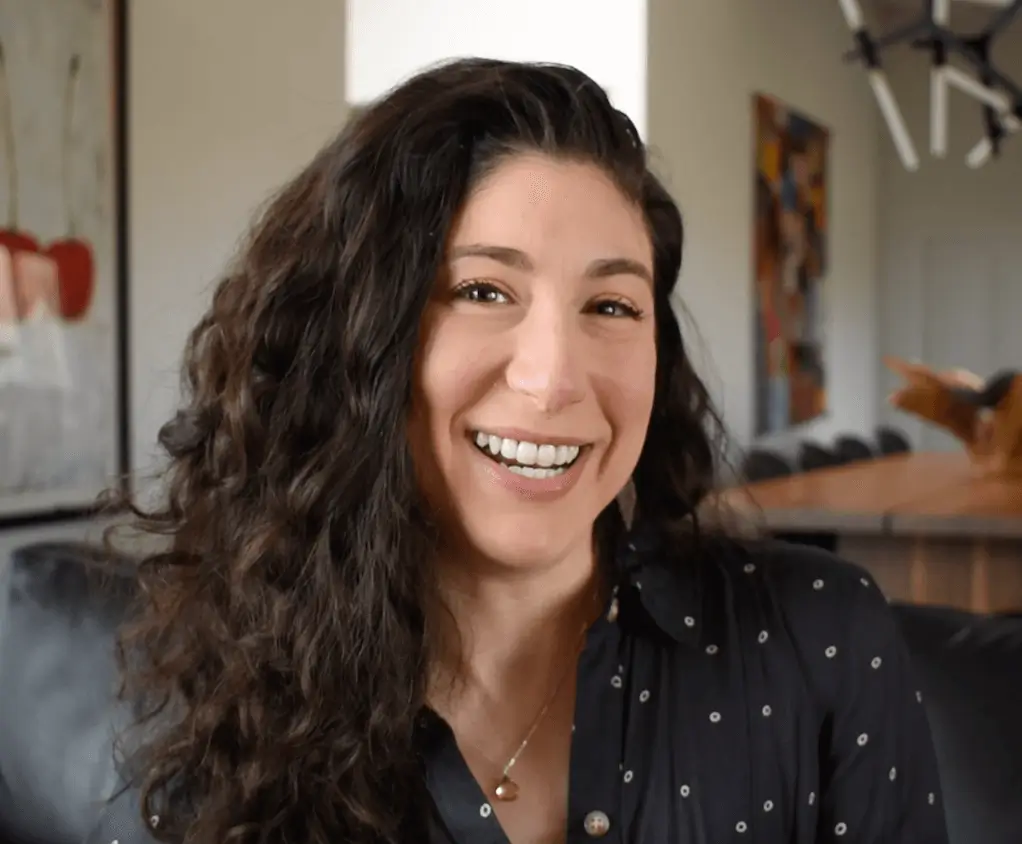 A woman with long curly brown hair smiles at the camera, wearing a dark shirt with small white dots. She is indoors, with abstract art and a dining table visible in the background.